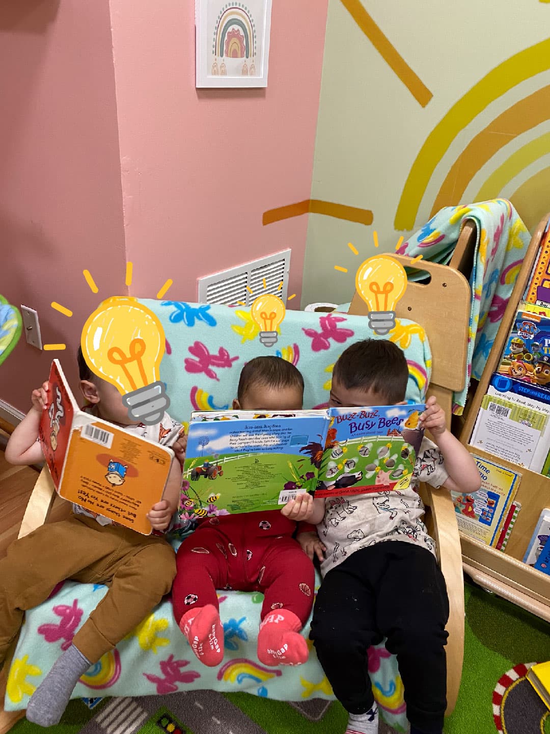 3 daycare kids sitting side by side reading. The toddler to the left is holding his book upside down, the other two are reading one book together. 